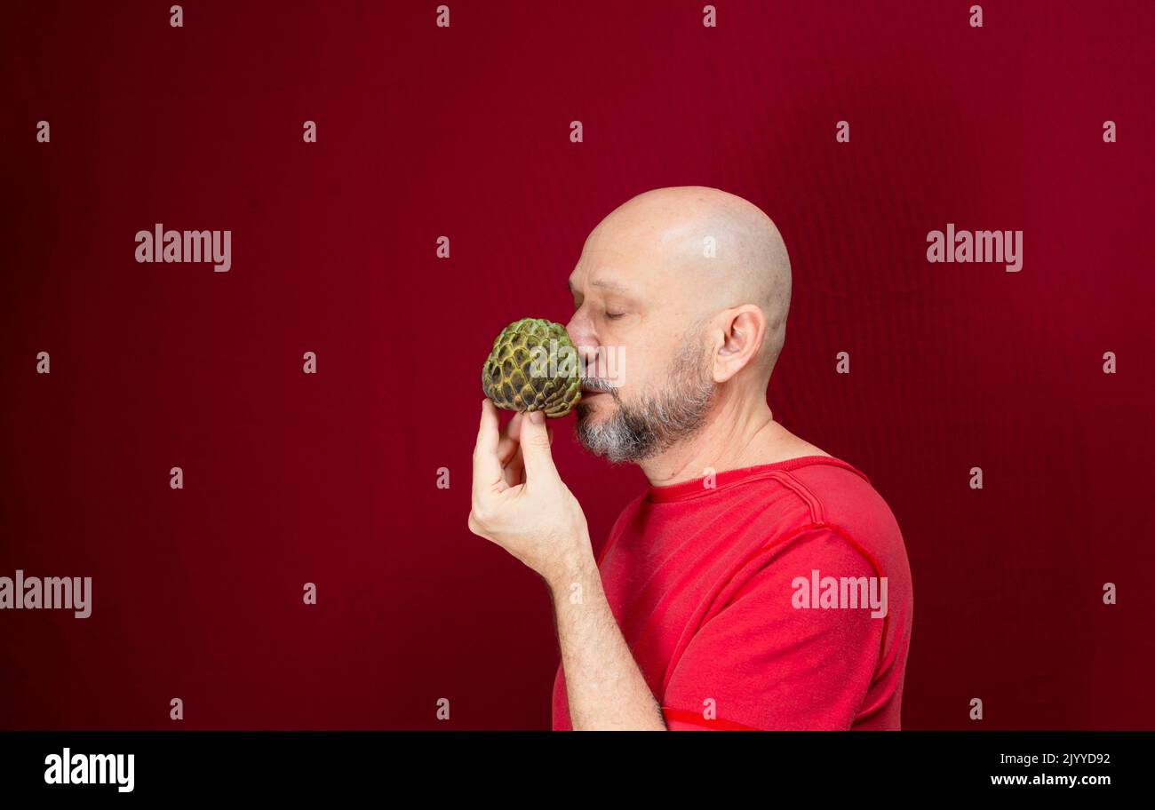 Young handsome man with beard, bald head and red shirt standing holding pinecone fruit against red background. Positive and healthy person. Stock Photo