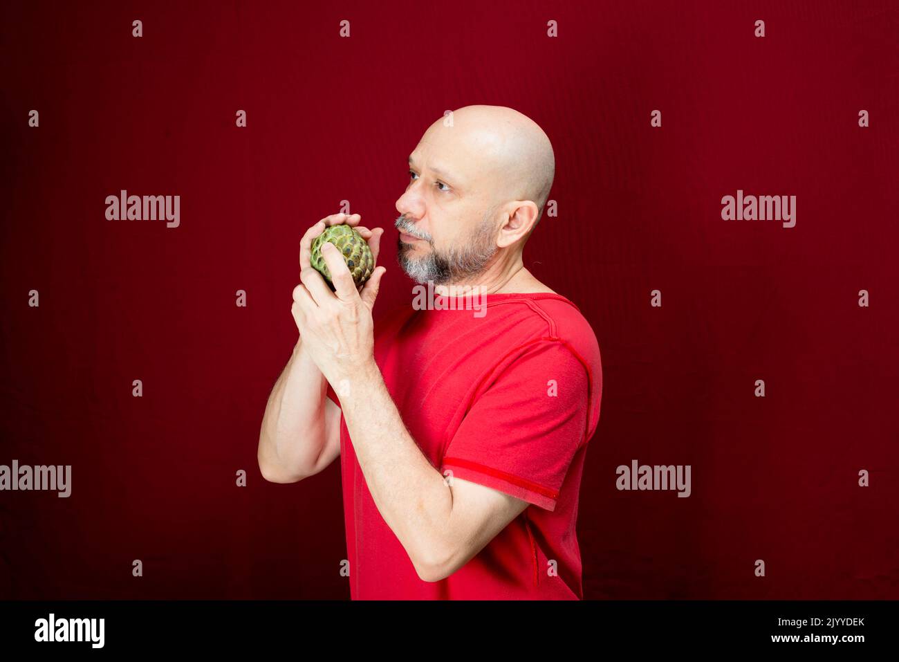 Young handsome man with beard, bald head and red shirt standing holding pinecone fruit against red background. Positive and healthy person. Stock Photo