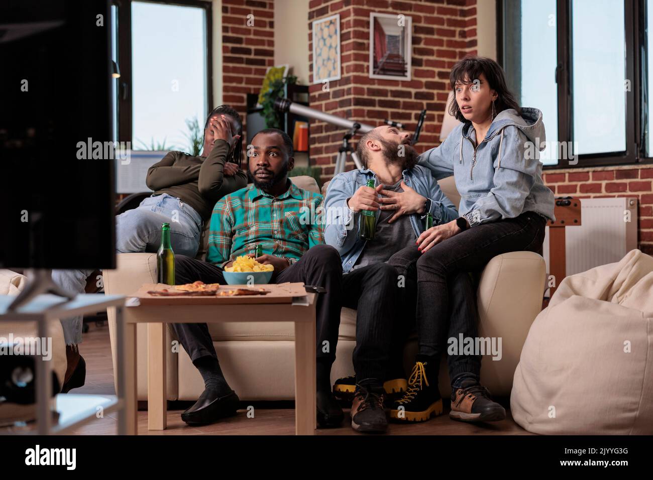 Group of friends getting scared and watching horror film on tv, having fun with beer bottles and snacks. People being frightened and looking at scary thriller movie, enjoying friendship gathering. Stock Photo