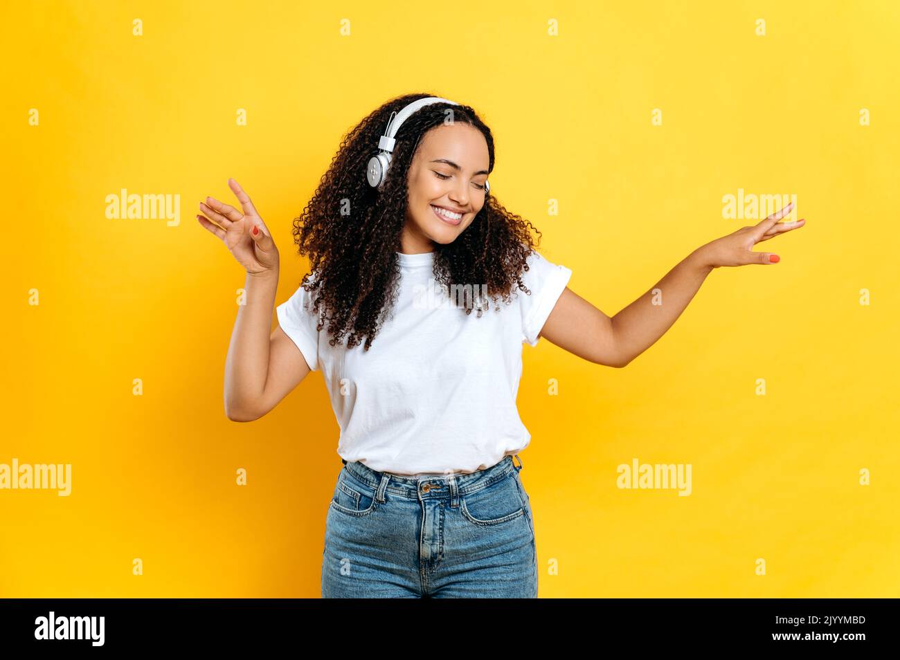 Cheerful lovely young brunette mixed race woman, in casual stylish wear, with headphones, listens her favorite music, dancing with eyes closed, relaxing, having fun on isolated orange background,smile Stock Photo