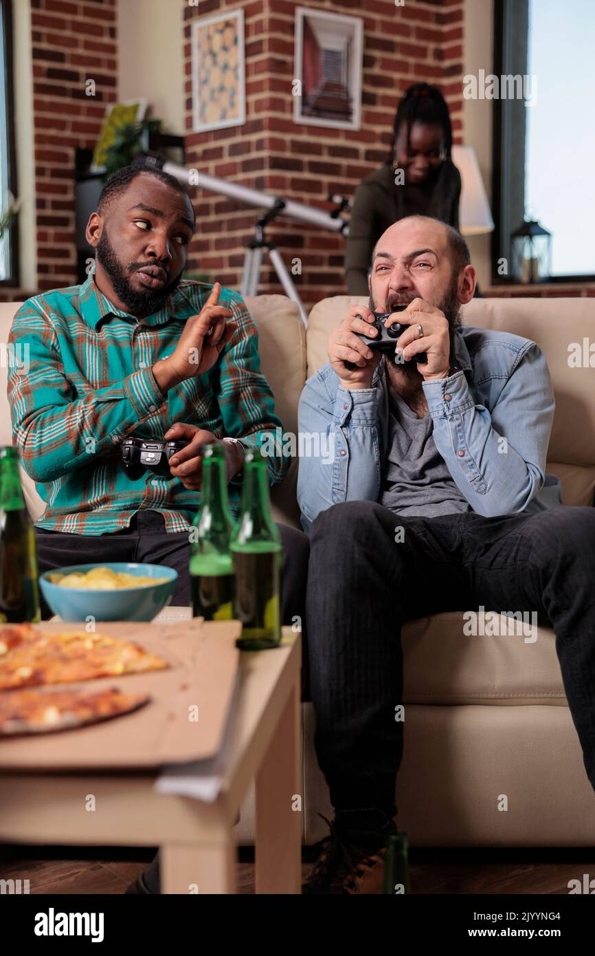 Team of friends losing video games competition on tv console, feeling sad and frustrated about lost game play. People gathering at house party with snacks and beer bottles for leisure. Stock Photo
