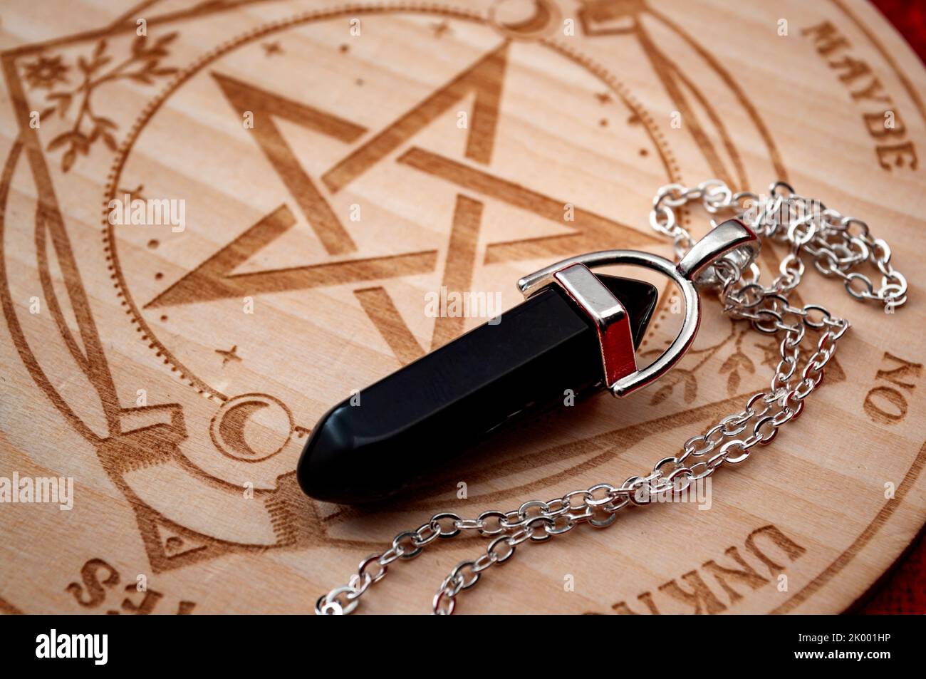 Dowsing pendulum with black gemstone next to hardwood divination chart on red velvet backgrounds concept for mystic magic spell, advanced esoteric sym Stock Photo