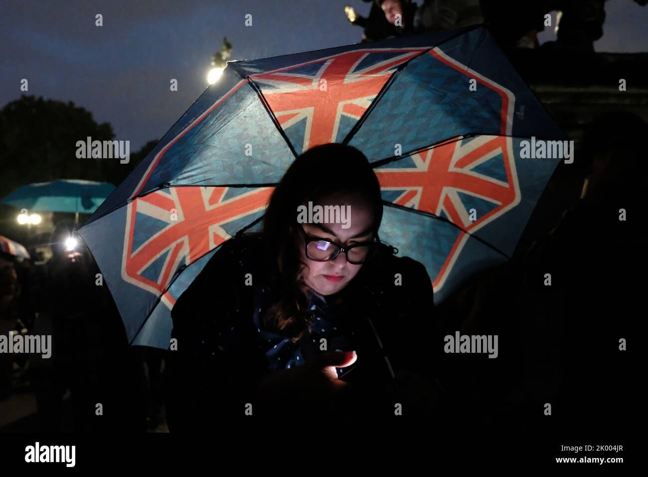 London, UK, 8th September, 2022. A woman looks at her phone whilst carrying a Union Jack covered umbrella. Members of the public paid their respects outside Her Majesty the Queen's London residence, Buckingham Palace until late into the night after her death was announced earlier during the evening. The country will now enter ten days of mourning as the monarch's 70 year reign comes to an end. Credit: Eleventh Hour Photography/Alamy Live News Stock Photo