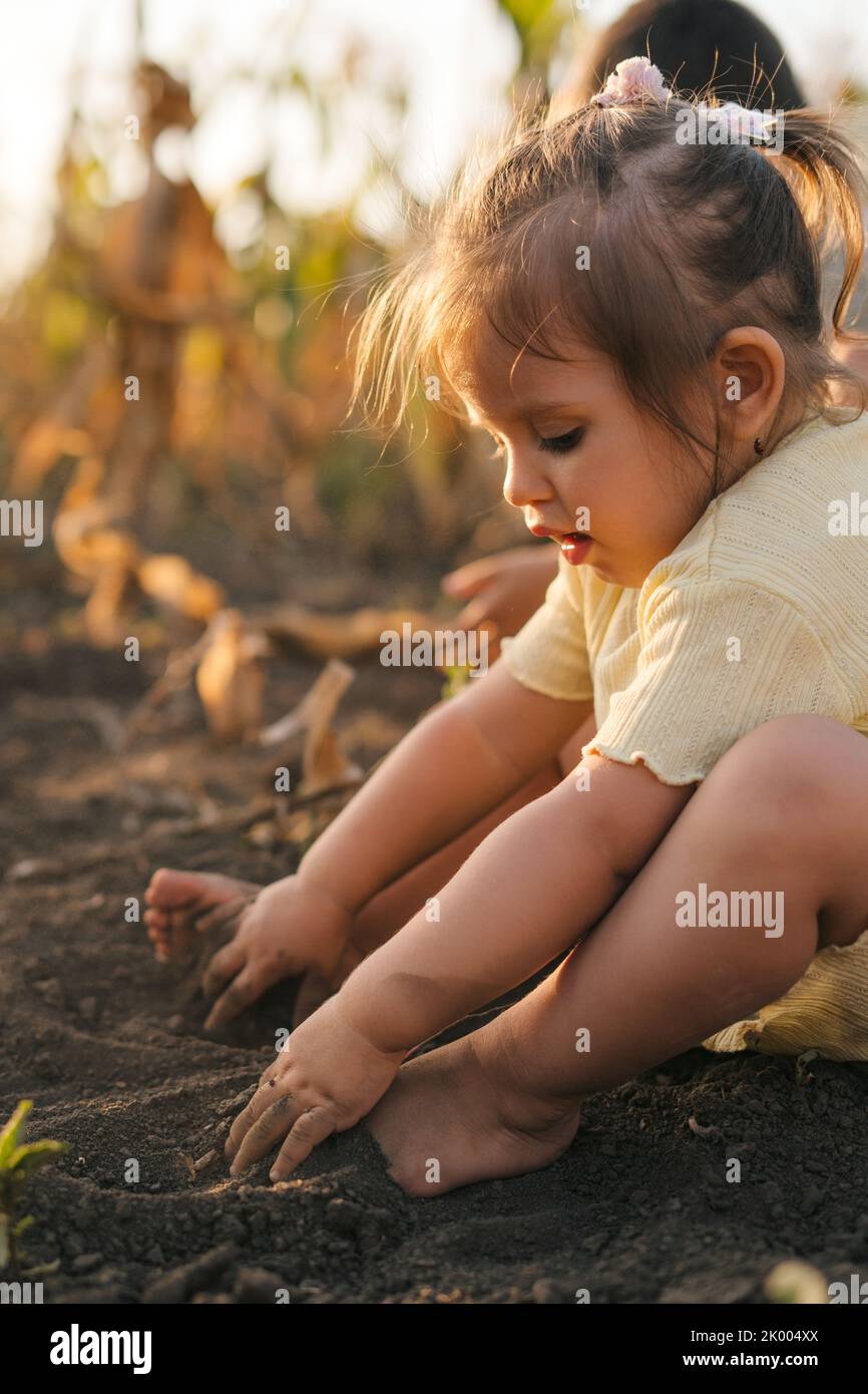 Baby girl sitting barefoot on the ground in the garden, digging with her hands in the ground, imitating parents planting seeds in the summer garden Stock Photo