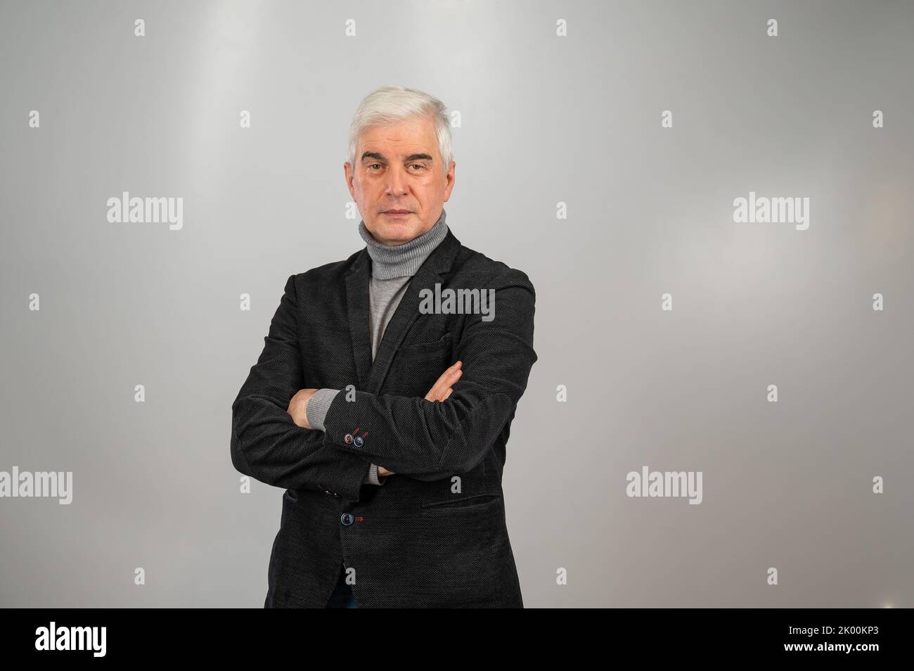 An elderly gray-haired man in a suit stands with his arms crossed over ...