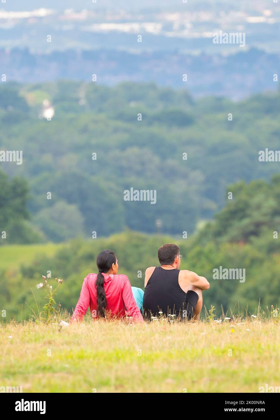Rear/back view of man and woman sitting isolated together on rural hillside looking at the view of a town in the distance. Stock Photo
