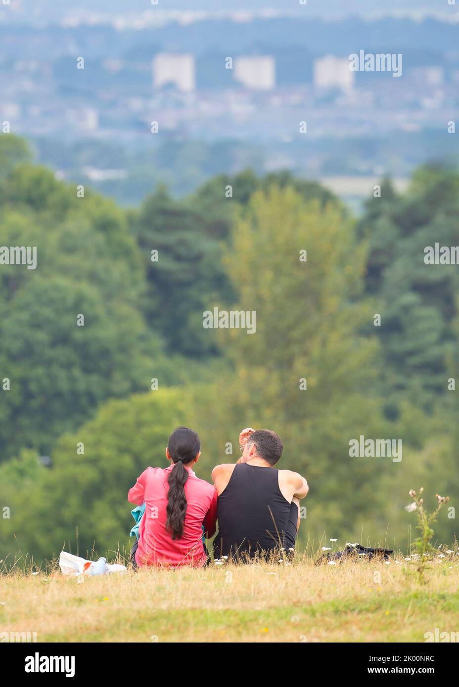 Rear/back view of man and woman sitting isolated together on rural UK hillside looking at the view of Birmingham city in the distance. Stock Photo