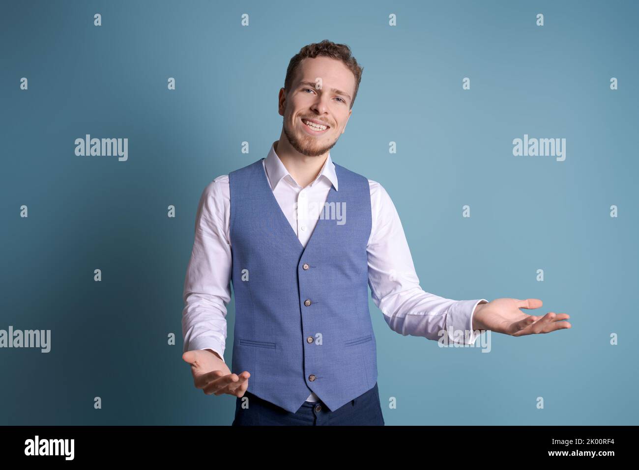 Portrait handsome excited man in business clothes smiling and showing hand gestures at the camera, isolated on blue background, young caucasian man with stubble on his face Stock Photo