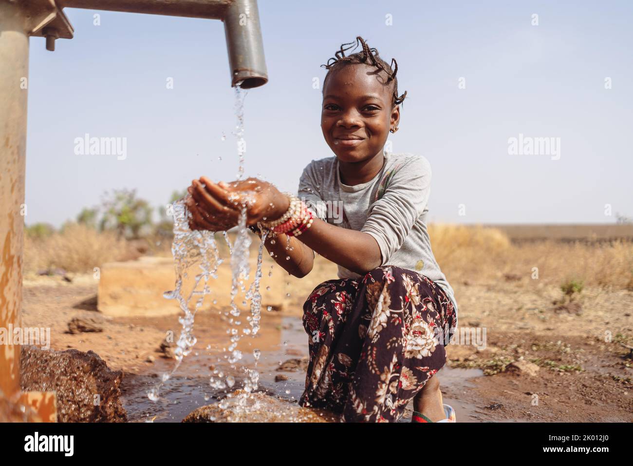 Thirsty African girl with funny braids, sitting in the middle of a puddle, happy to collect the abundant water that flows from the village fountain; c Stock Photo