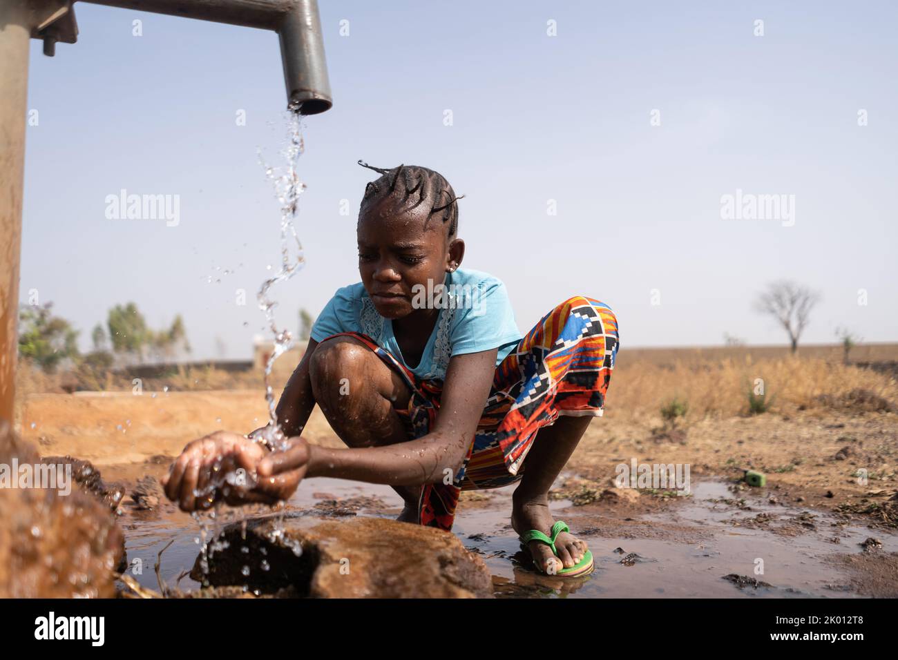A young African girl casually crouches in front of a water pump and washes roughly, which is symbolic of the lack of tap water and washing facilities Stock Photo
