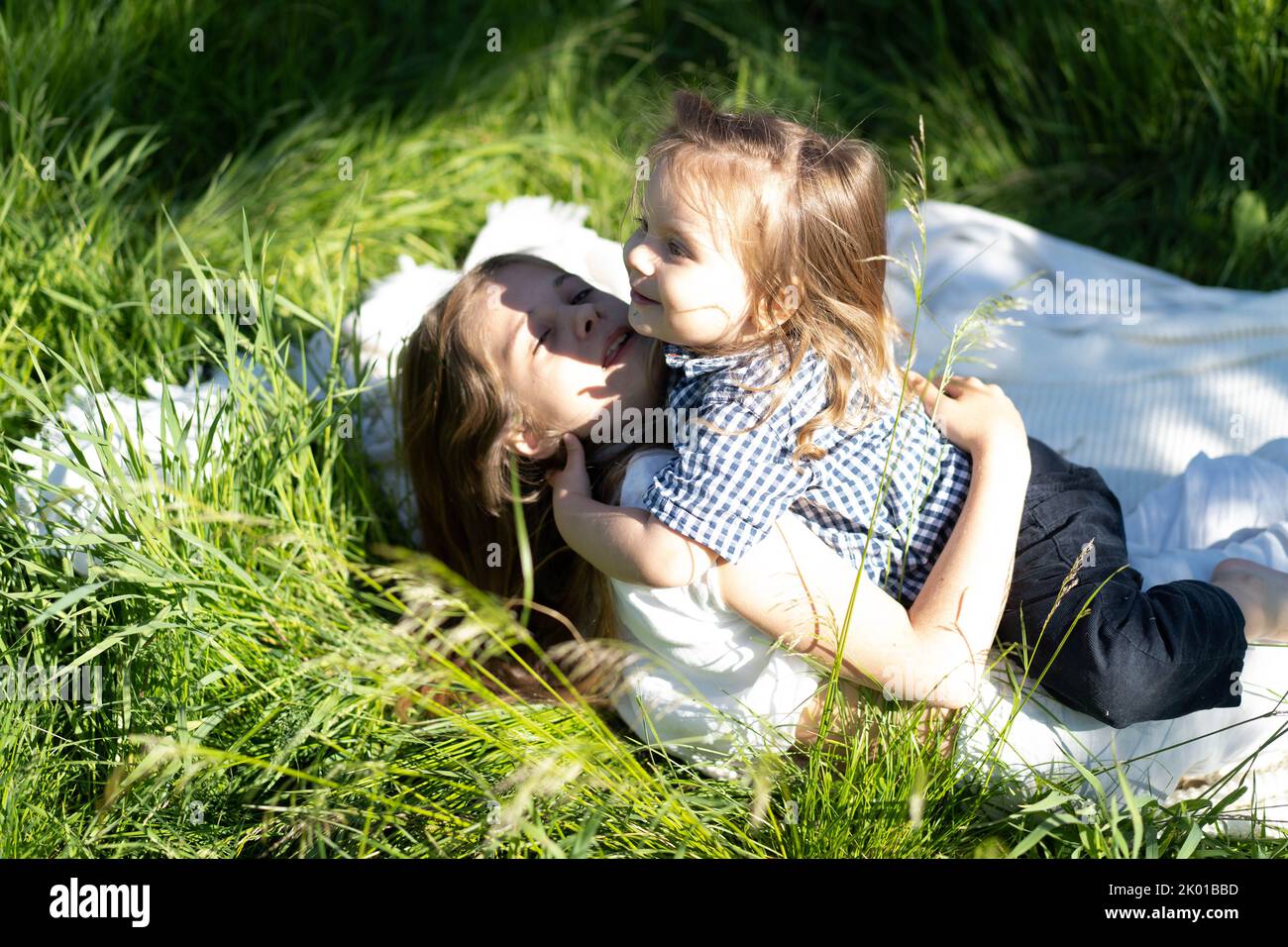 Brother and sister happily hug and laugh, lying on the green grass. The sun is shining, the children are happy carefree. The concept of a happy family life and bonding. Copy space. Stock Photo