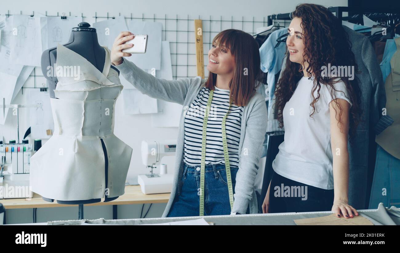 Two creative female designers are making selfie with smart phone while standing beside tailoring dummy in modern workplace. Attractive women are smiling and posing. Stock Photo