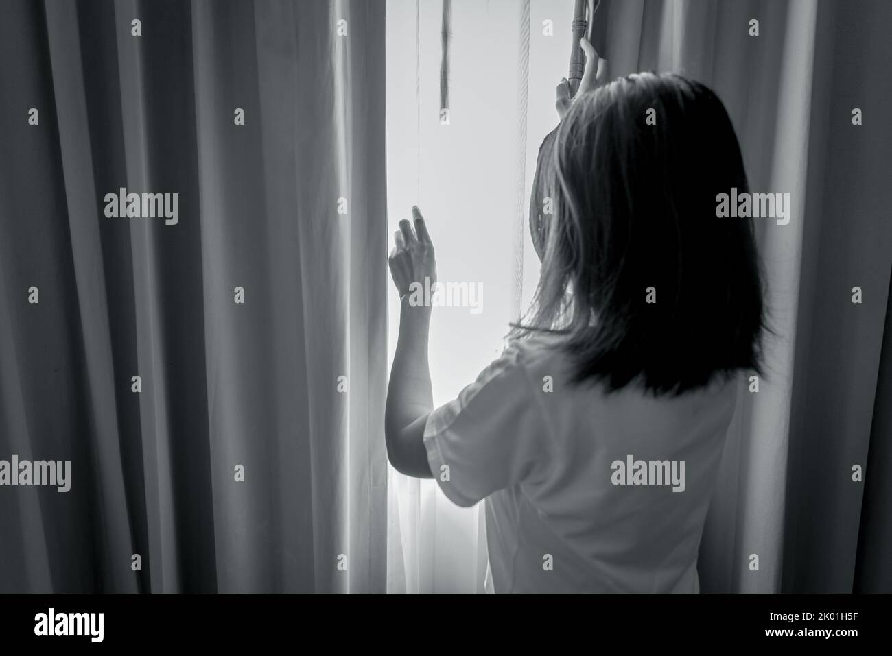 Rear view of woman holding curtain and looking out of the bedroom. Depressed and stressed woman. World mental health day concept. Mental health Stock Photo