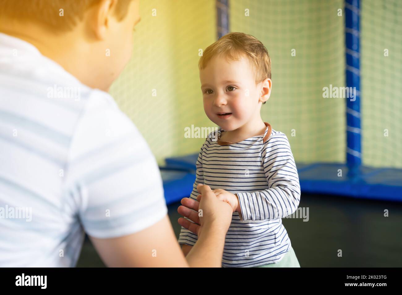 Dad holds the hand of a boy jumping on a trampoline. Family time. Boy of one and a half years. Active entertainment. Toddler. Focus on the boy's eyes Stock Photo