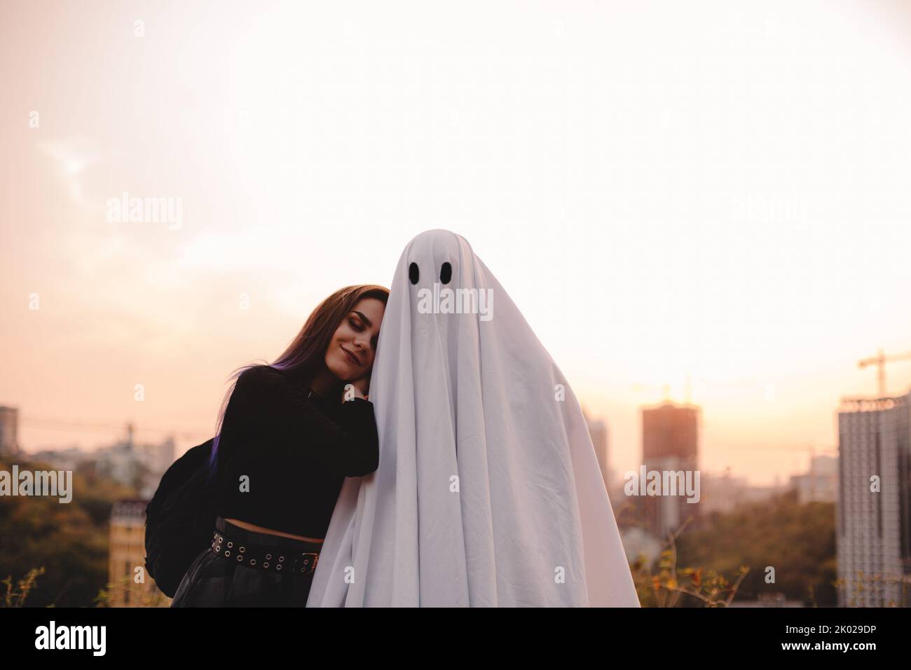 Happy young woman leaning on ghost standing against evening city during Halloween at sunset Stock Photo