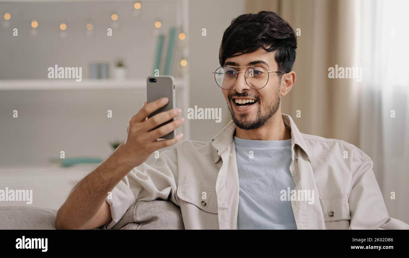 Portrait indoor male arabic guy unshaven man with glasses sitting on couch at home chatting online using mobile phone reads news scrolling browsing Stock Photo