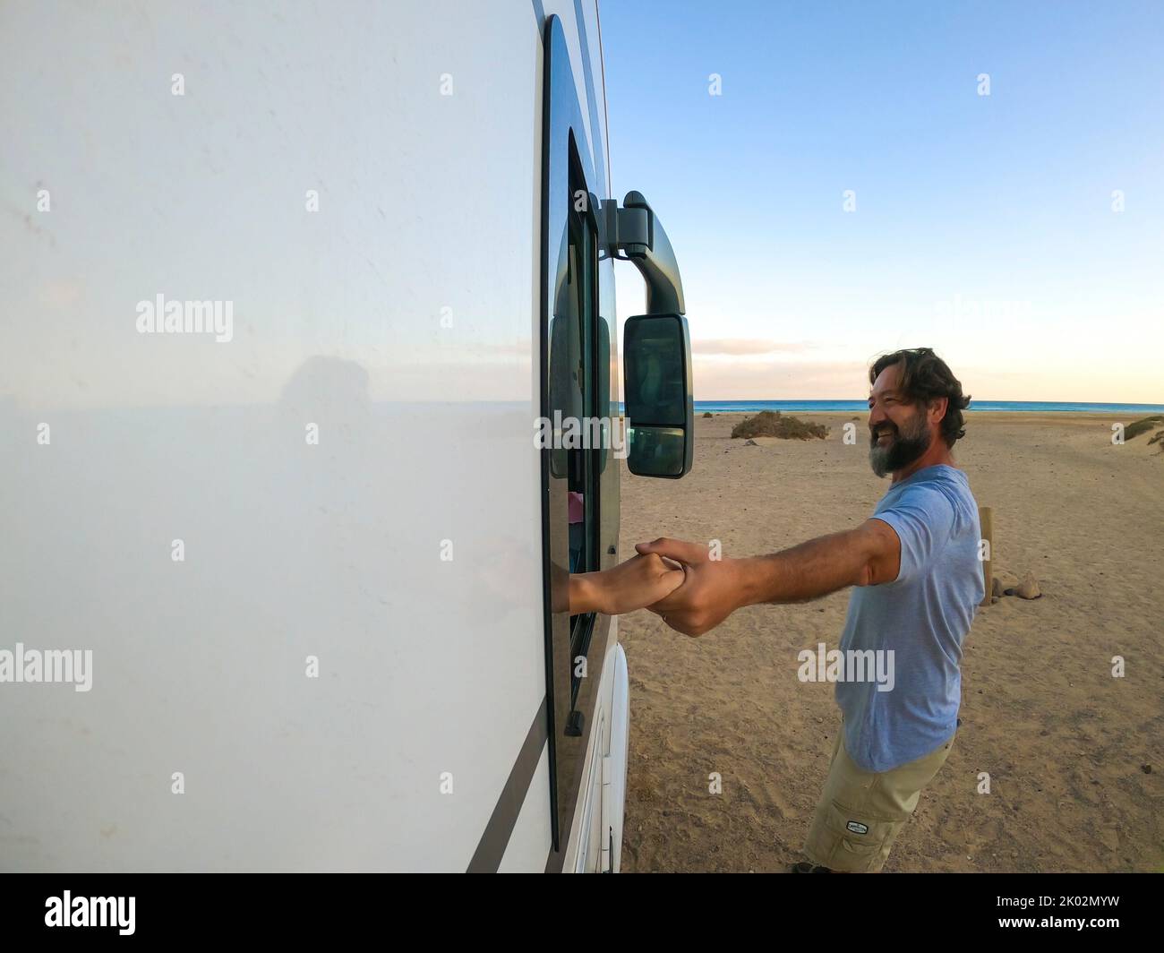 Outside view of couple holding hands. Woman inside a camper van and man outside smiling. Travel for holiday vacation lifestyle concept. beach and ocean in background Stock Photo