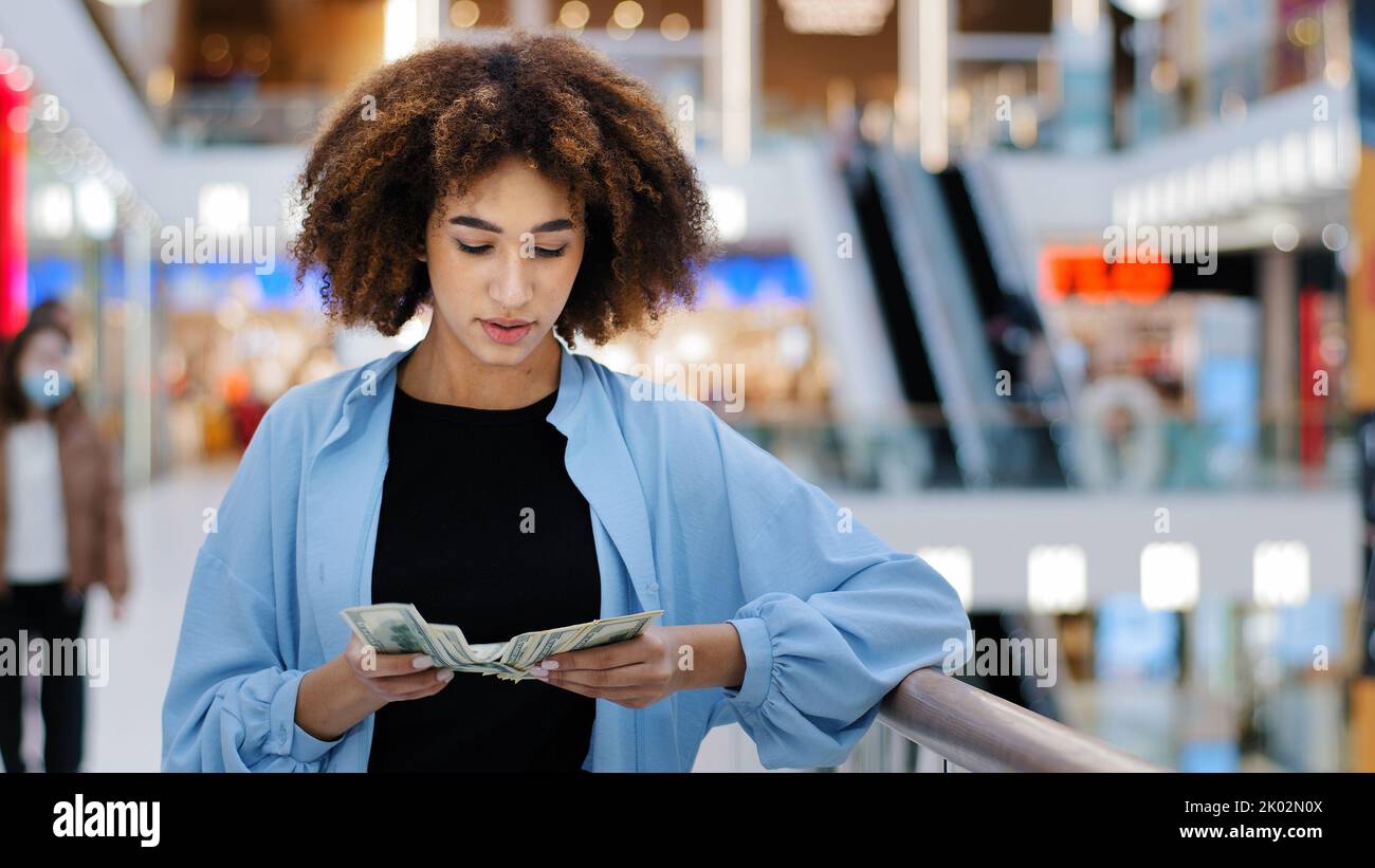 Millennial african american girl shopper consumer client woman standing in shopping business center counting money cash dollars banknotes earning Stock Photo