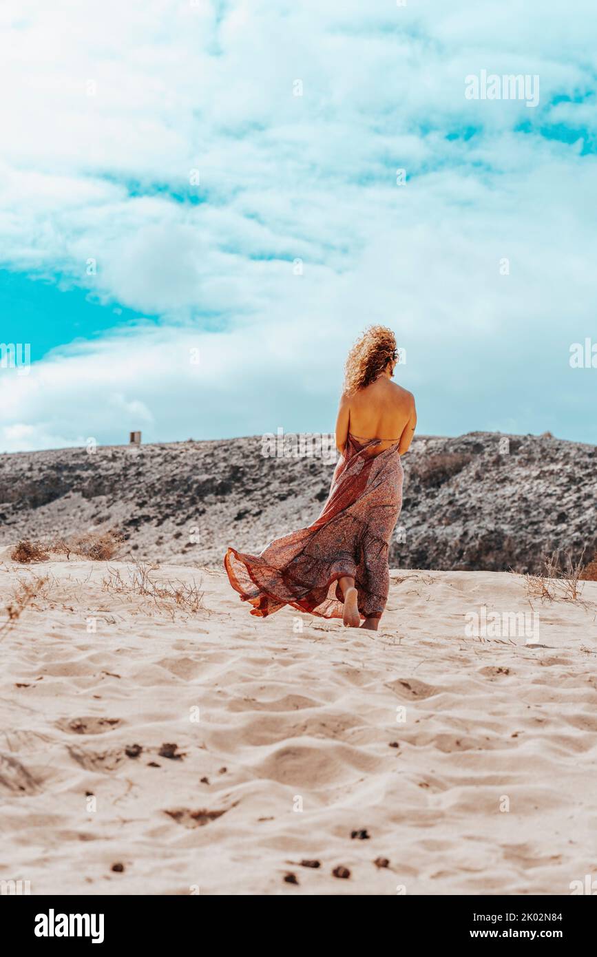 Standing woman with boho elegant dress in front of a desert mountains and blue sky with clouds. Travel and serene lifestyle people. Female with nude back walk barefoot on the sand in summer Stock Photo