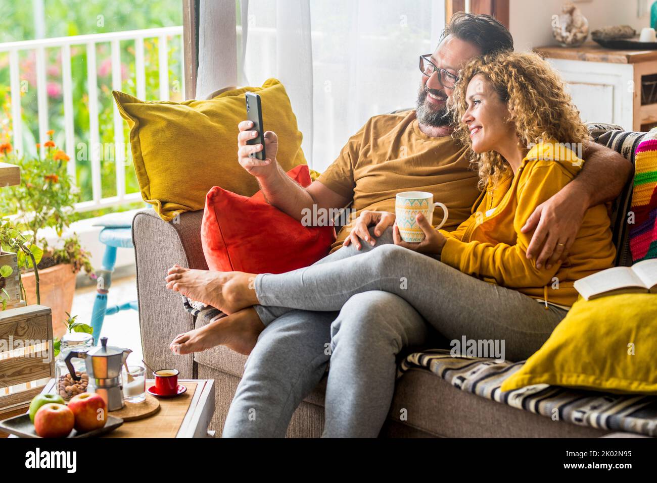 Caucasian couple doing video call with mobile phone at home sitting on the sofa. Man and woman enjoying technology wireless connection during morning breakfast and have fun calling friends Stock Photo