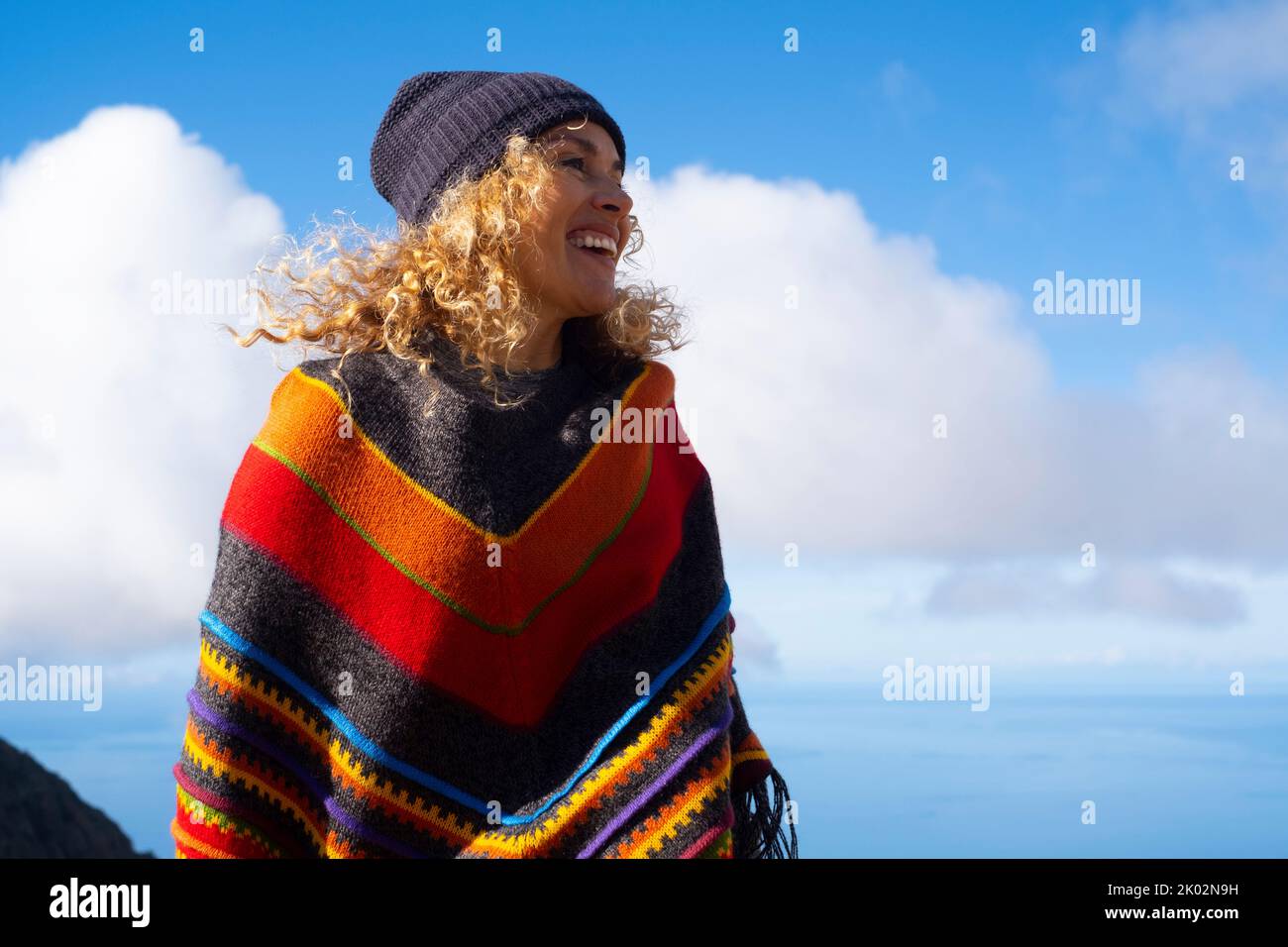 Colorful poncho clothes on happy pretty adult woman enjoying outdoor leisure activity against a blue sky background. Happy and cheerful people female portrait with winter hat Stock Photo