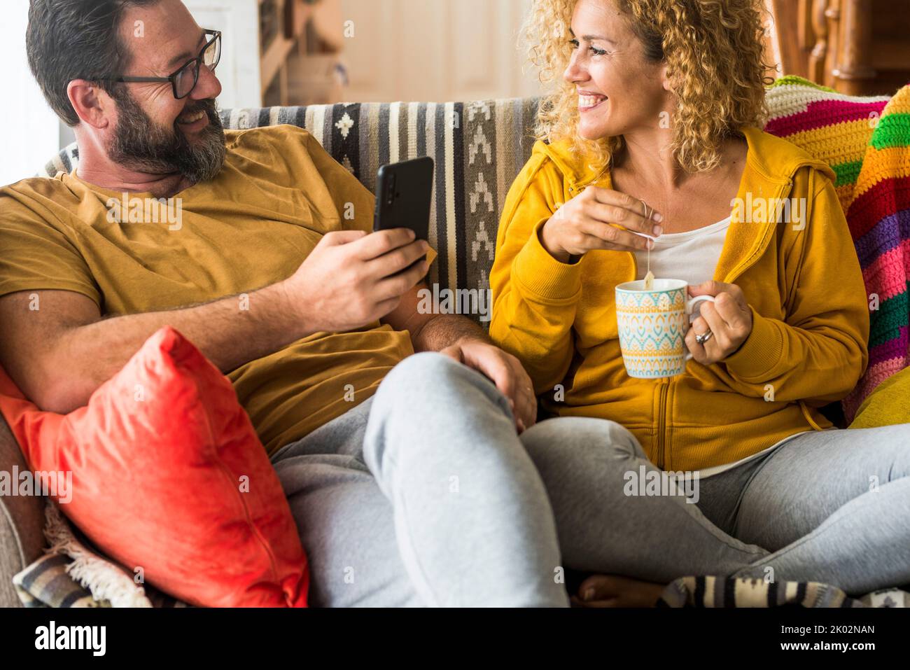 Man and woman smile and enjoy leisure activity at home in morning breakfast time together. People sitting on the sofa in apartment for real life. Couple use phone and drink tea having fun on the couch Stock Photo