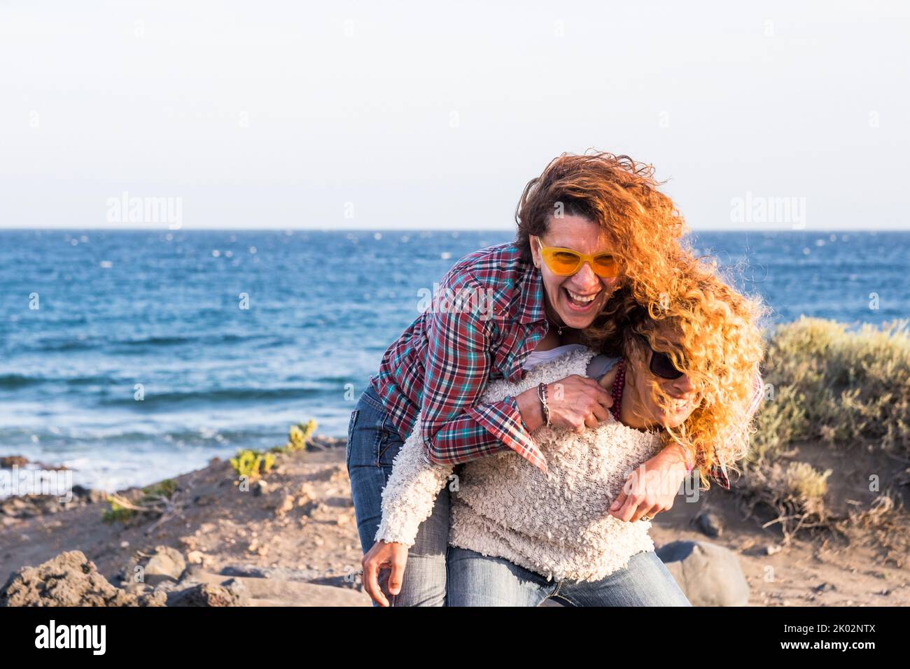 Couple of young adult woman having fun carrying in piggyback and laughing a lot. Curly hair female people enjoy leisure activity outdoor with ocean in background. Summer holiday vacation lifestyle Stock Photo
