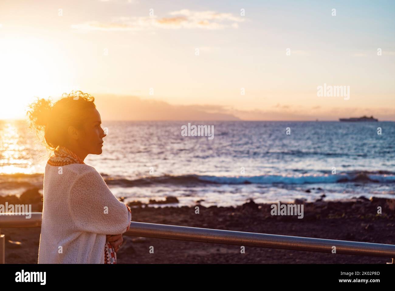 Side view of young adult woman admiring and enjoying golden sunset at the beach looking at the sea. Female people and nature outdoor leisure activity. Thoughtful lady and travel life Stock Photo