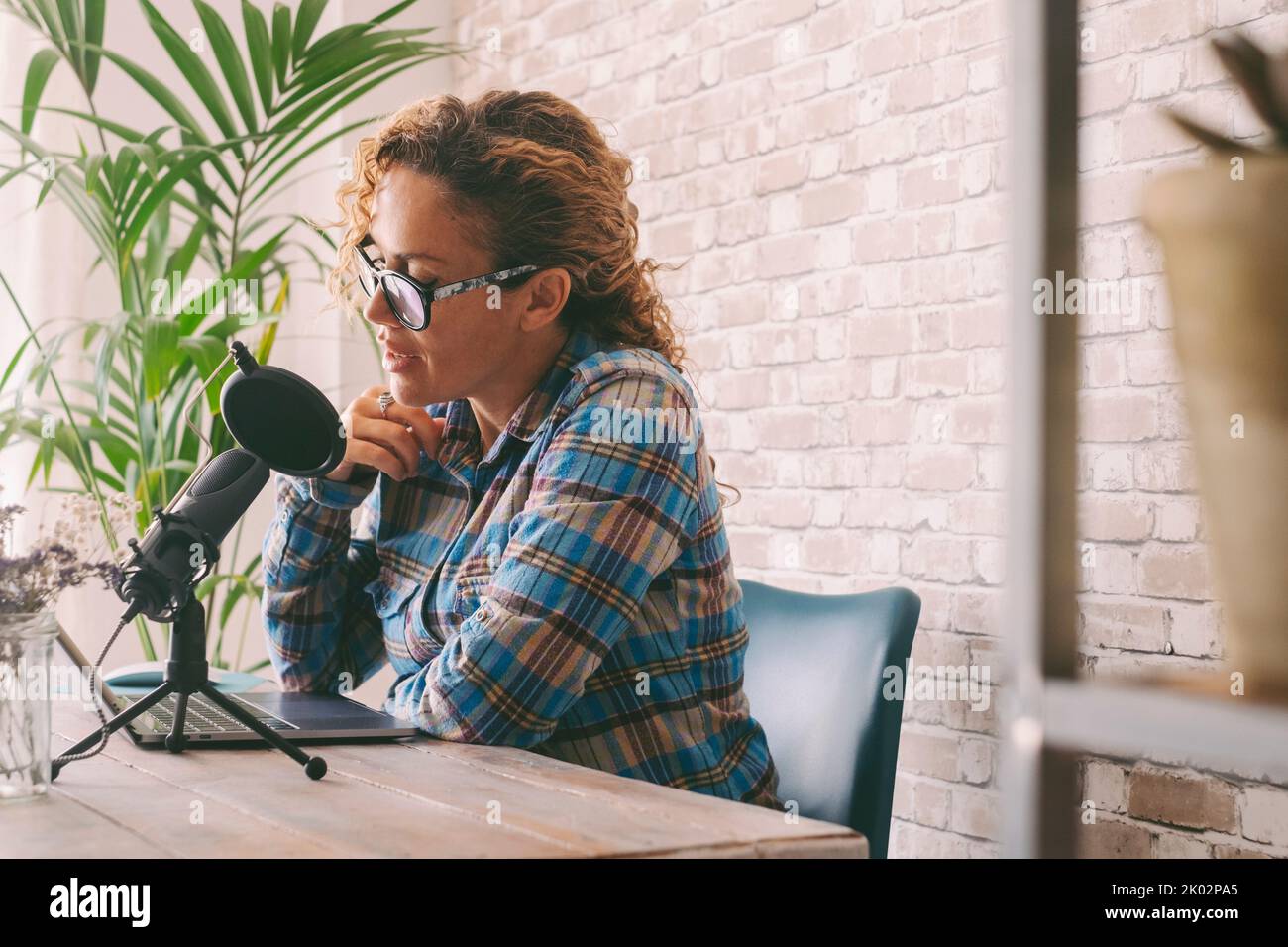 Content creator adult woman recording a podcast interview using microphone and laptop sitting at the desk. Home office workstation and smart working modern people lifestyle. Online radio computer job Stock Photo