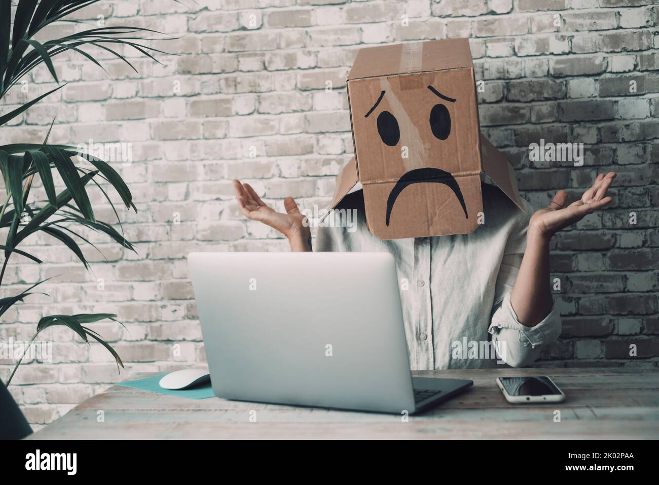 Modern worker with carton sad box on head open arms with desolation gesture in front of a laptop. Online crypto smart working business job activity. Concept of fail and lost work. Bad day office Stock Photo