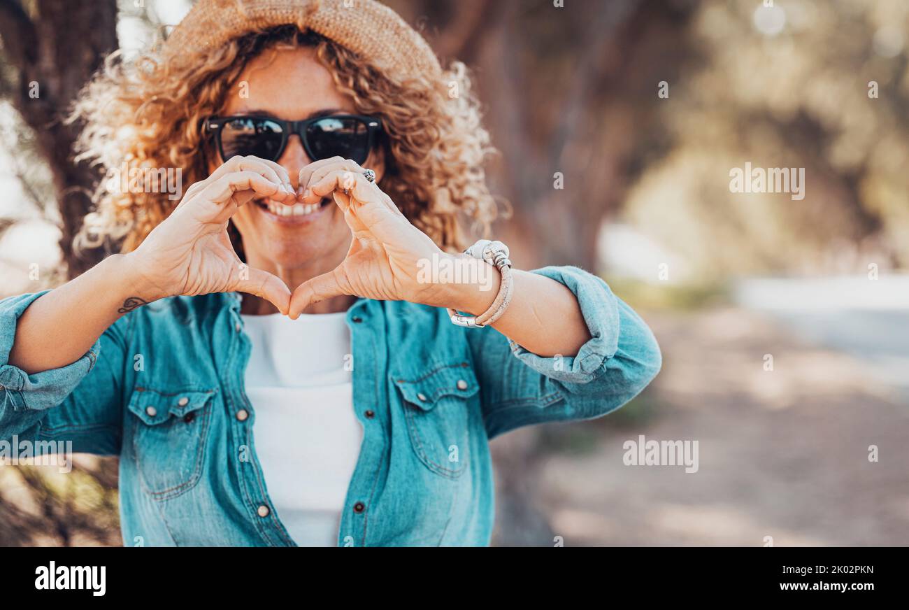 Pretty trendy young woman smile and have fun doing heart gesture sign symbol with hands to the camera. Outdoor leisure activity and trees in background. Happy people in tourism and travel lifestyle Stock Photo