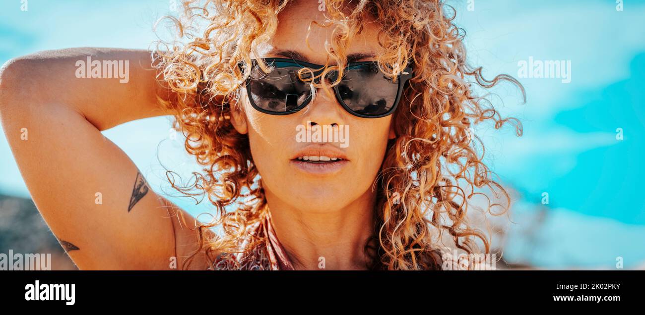 Summer portrait of beautiful young adult woman with sunglasses and healthy blonde long curly hair looking at camera. Attractive brunette with blue sky in background. One cute female people lifestyle Stock Photo