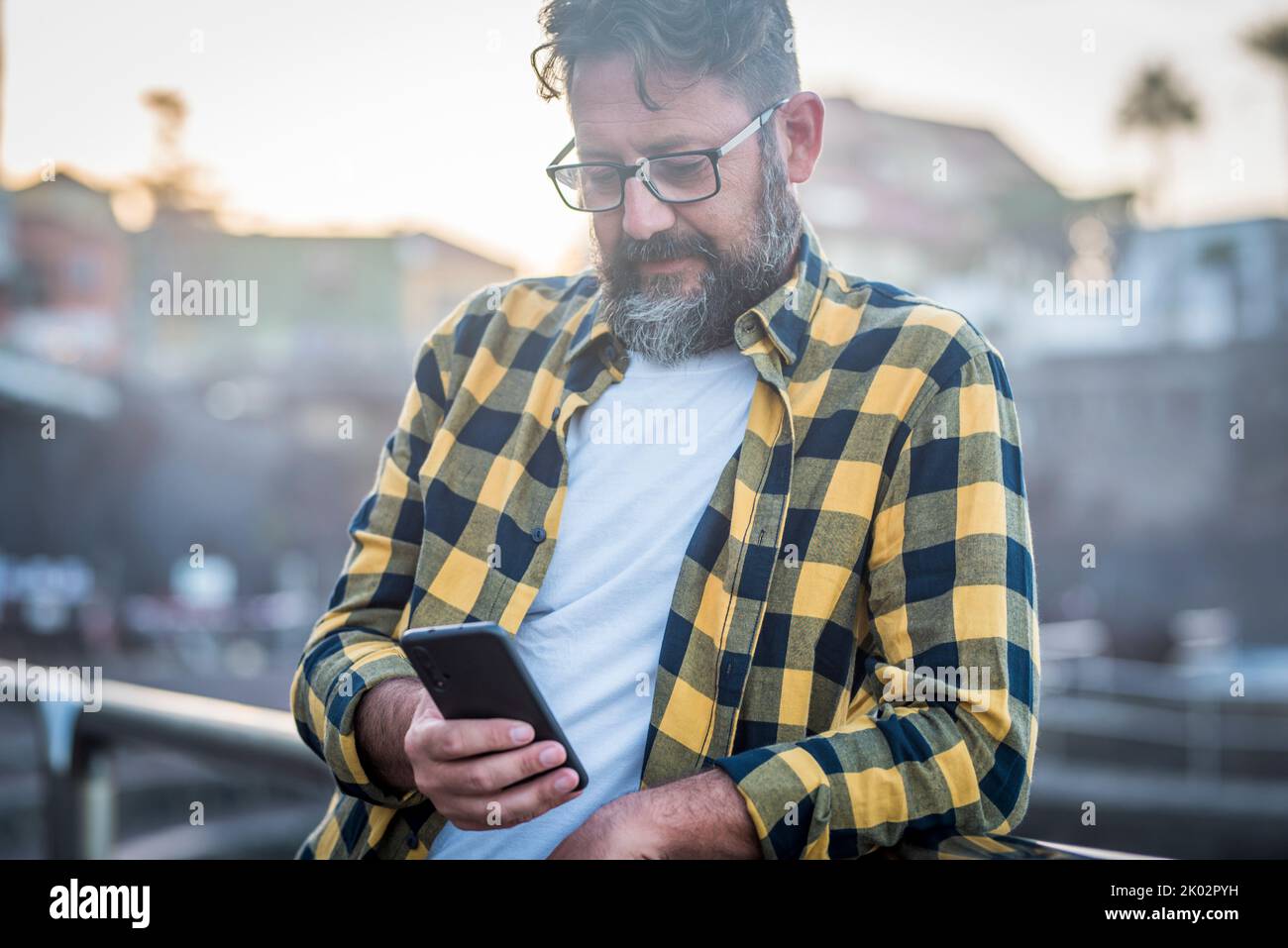 Mature bearded man use mobile phone with internet wireless connection wearing glasses. Adult male people chatting with smartphone outside. Sunlight in background. Leisure activity Stock Photo