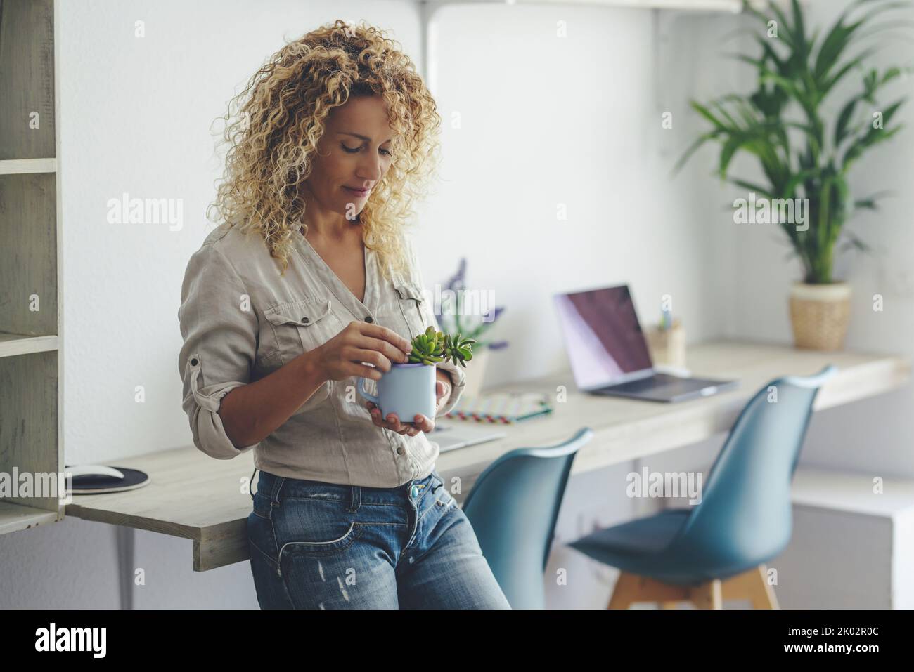 Pretty young mature lady woman enjoy indoor leisure activity at home standing and caring a plant. Laptop computer in background as alternative office workstation. Modern cute lady in casual wear smile Stock Photo