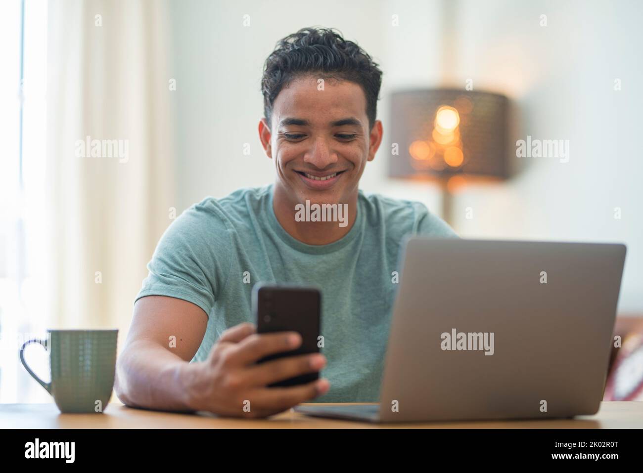 Young black boy using phone and computer connection at home. Happy man reading notification on mobile cellular while work on laptop in smart working. Modern Z generation people using web Stock Photo