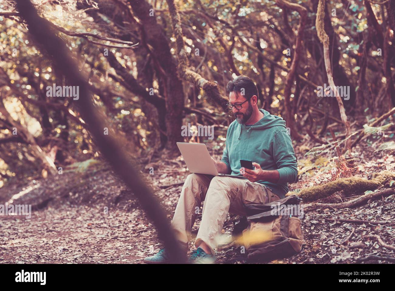 Man sit down in the outdoors nature forest and work with mobile phone and laptop computer with wireless roaming connection. Modern people enjoy alternative work place. Digital nomad travel Stock Photo