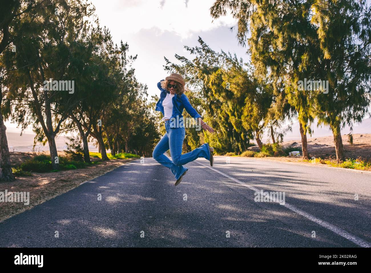 Young adult woman jumping happy in the middle of long straight asphalt road. Concept of travel people and freedom happiness emotions. Casual female have fun and enjoy road trip vacation Stock Photo