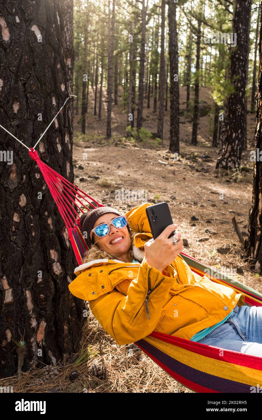View of woman enjoying freedom and independence lay down on colorful hammock in the forest woods nature park and using modern online phone to call and work in smart working activity Stock Photo