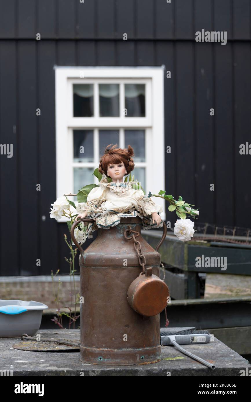 Milk can with doll, white roses, behind it facade of a wooden house, Marken peninsula, Waterland, Noord-Holland, Netherlands Stock Photo