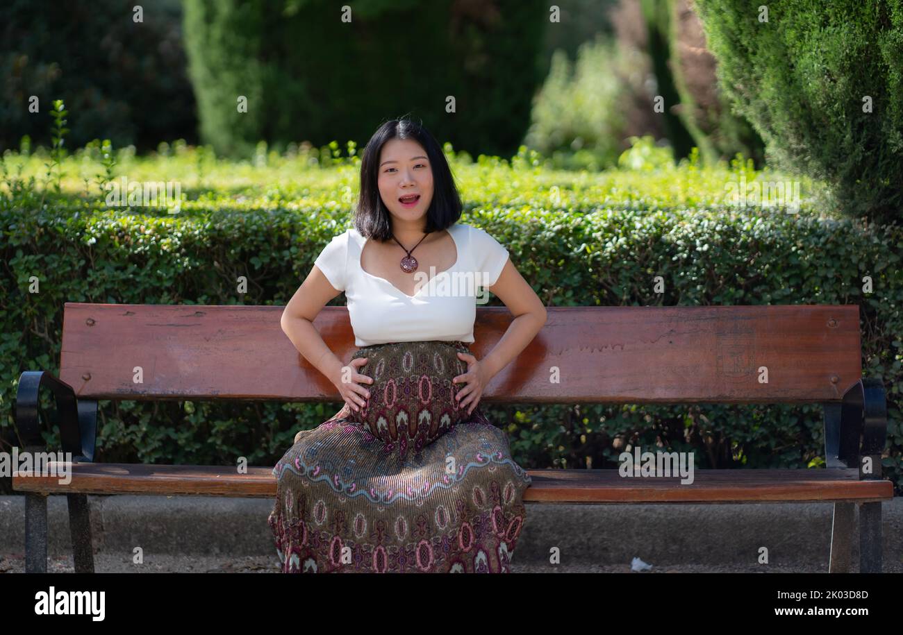 young happy and beautiful Asian Japanese woman pregnant showing her belly proud and cheerful smiling at city park bench happy in pregnancy and materni Stock Photo