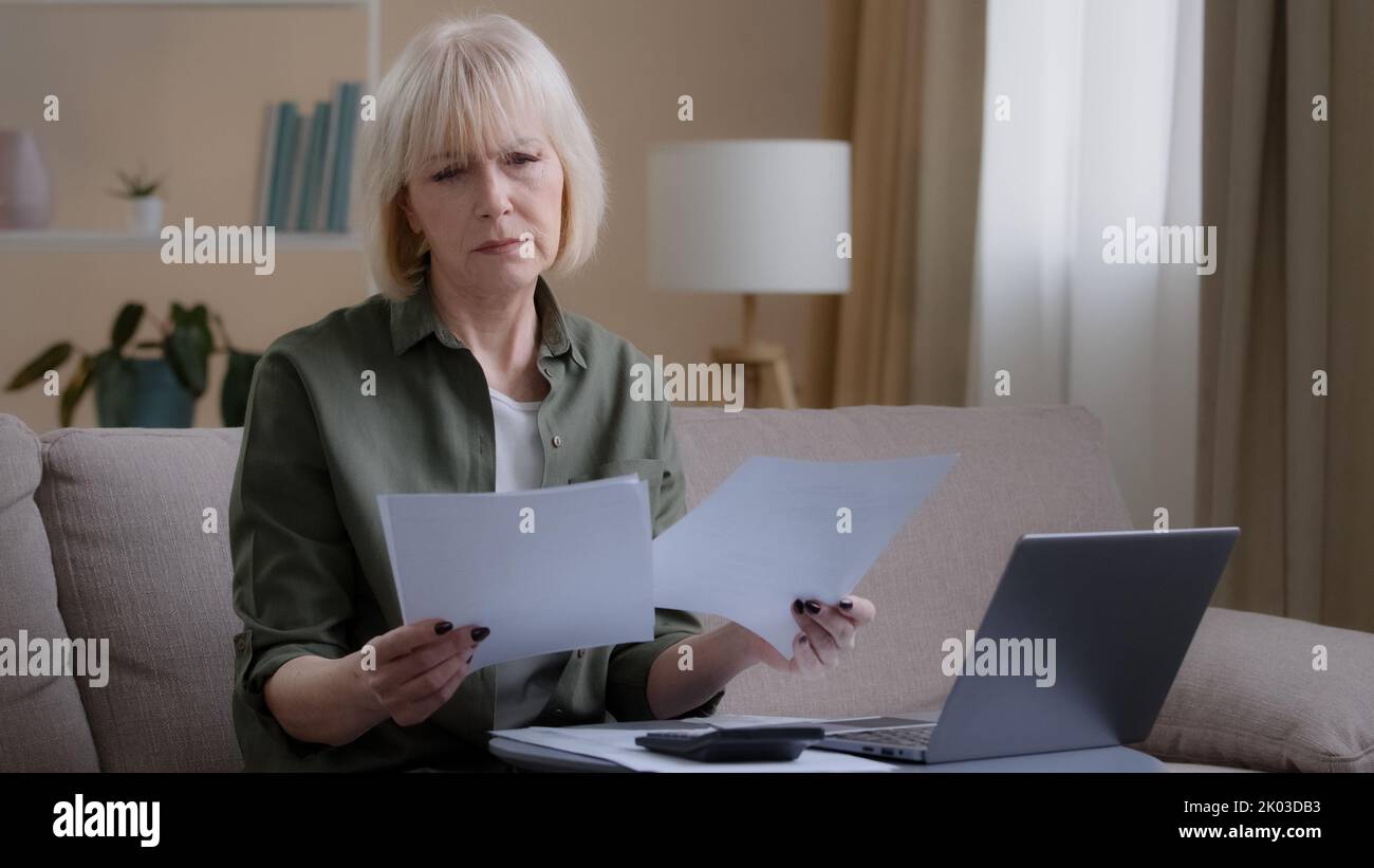 Caucasian old sad homeowner housewife makes woman budget checks monthly income by checking paper notes using app laptop. Middle-aged businesswoman Stock Photo