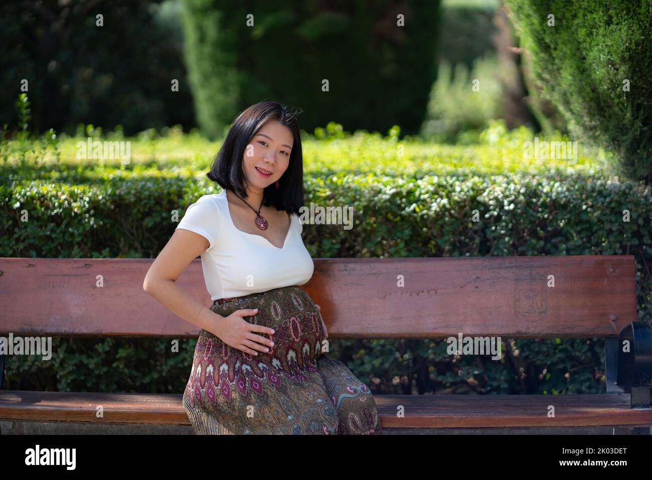 young happy and beautiful Asian Japanese woman pregnant showing her belly proud and cheerful smiling at city park bench happy in pregnancy and materni Stock Photo