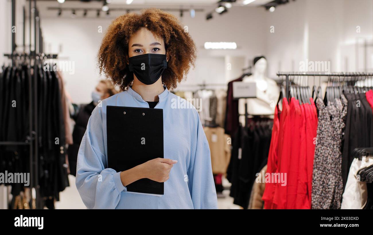 Black Friday. African american welcoming woman saleswoman girl consultant in medical mask boutique store worker standing in shopping mall inviting to Stock Photo