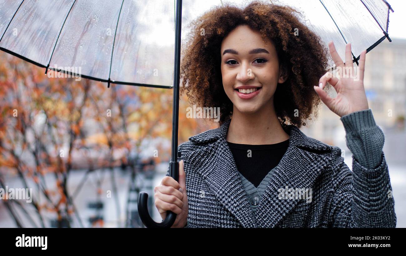 Beautiful happy successful lady curly african american woman girl with transparent umbrella stands in autumn city in rain looking at camera smiling Stock Photo
