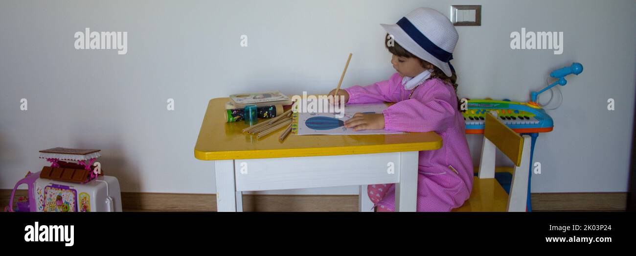 Adorable little girl with pink hat and apron playing with pencil colors in a notebook. Back to school and first day of kindergarten. Horizontal banner Stock Photo
