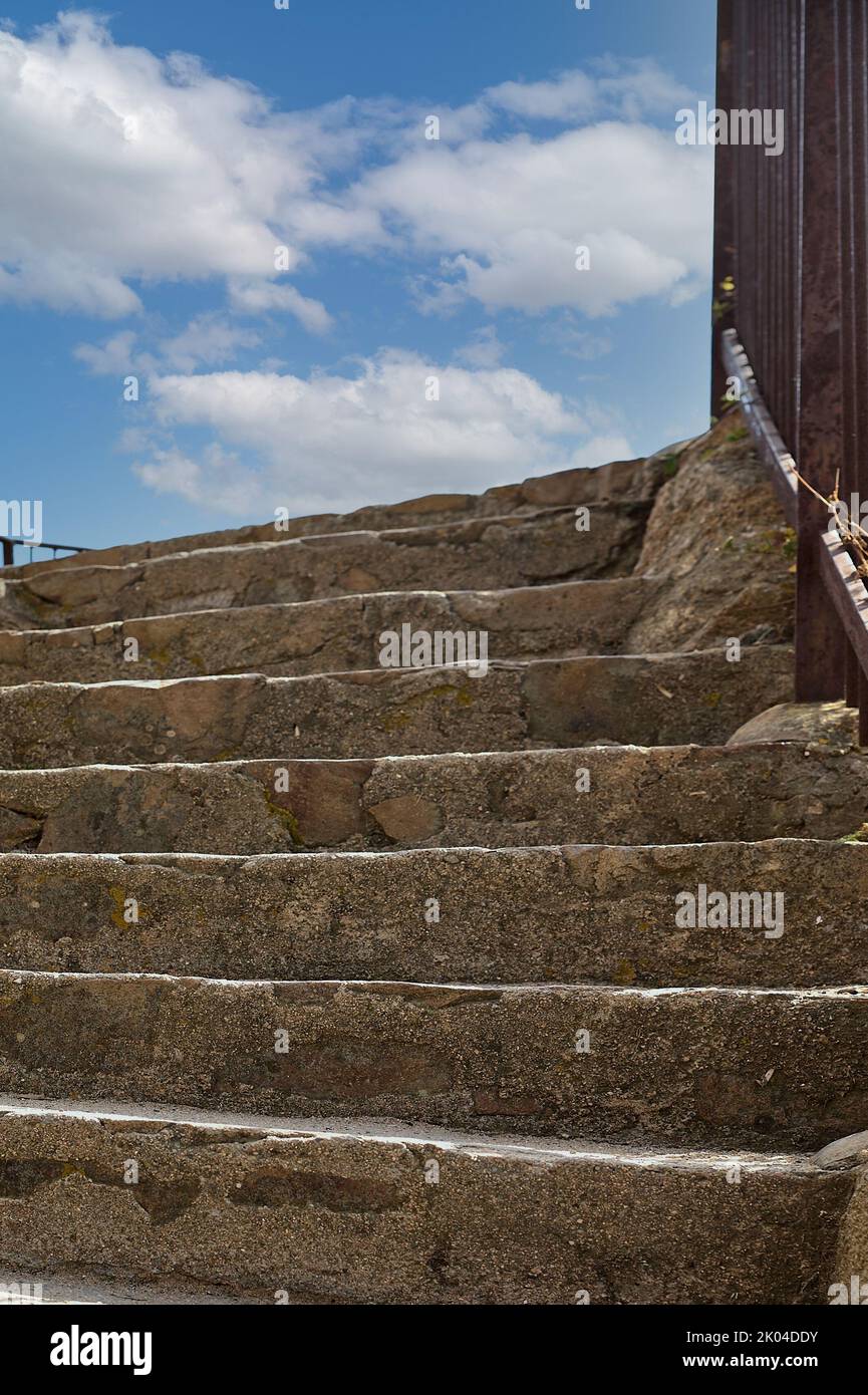 Stone staircase with iron railing on one side that goes up and ends in a blue sky with very white clouds. Stock Photo