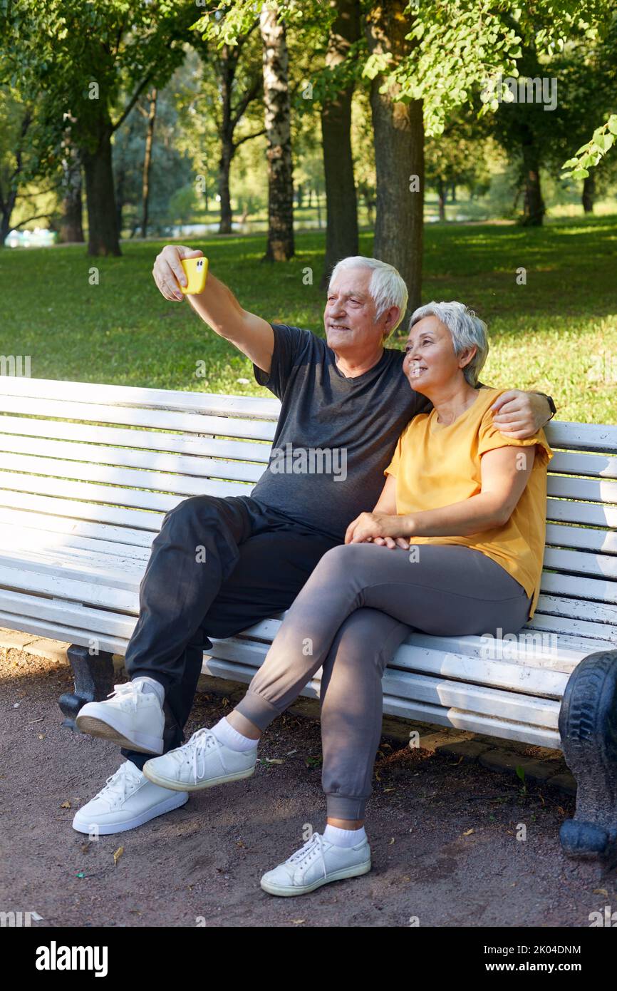 Happy senior spouses take selfie picture sit on bench in summer park. Grey-haired couple look at cell phone screen make videocall outdoor use modern t Stock Photo