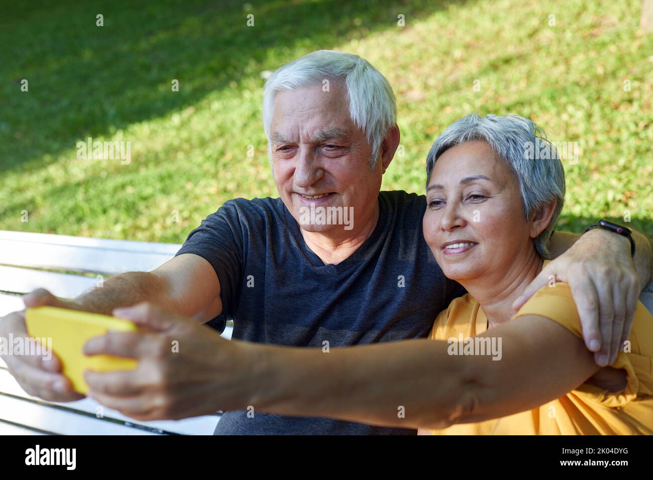 Happy senior spouses take selfie picture sit on bench in summer park. Grey-haired couple look at cell phone screen make videocall outdoors use modern Stock Photo