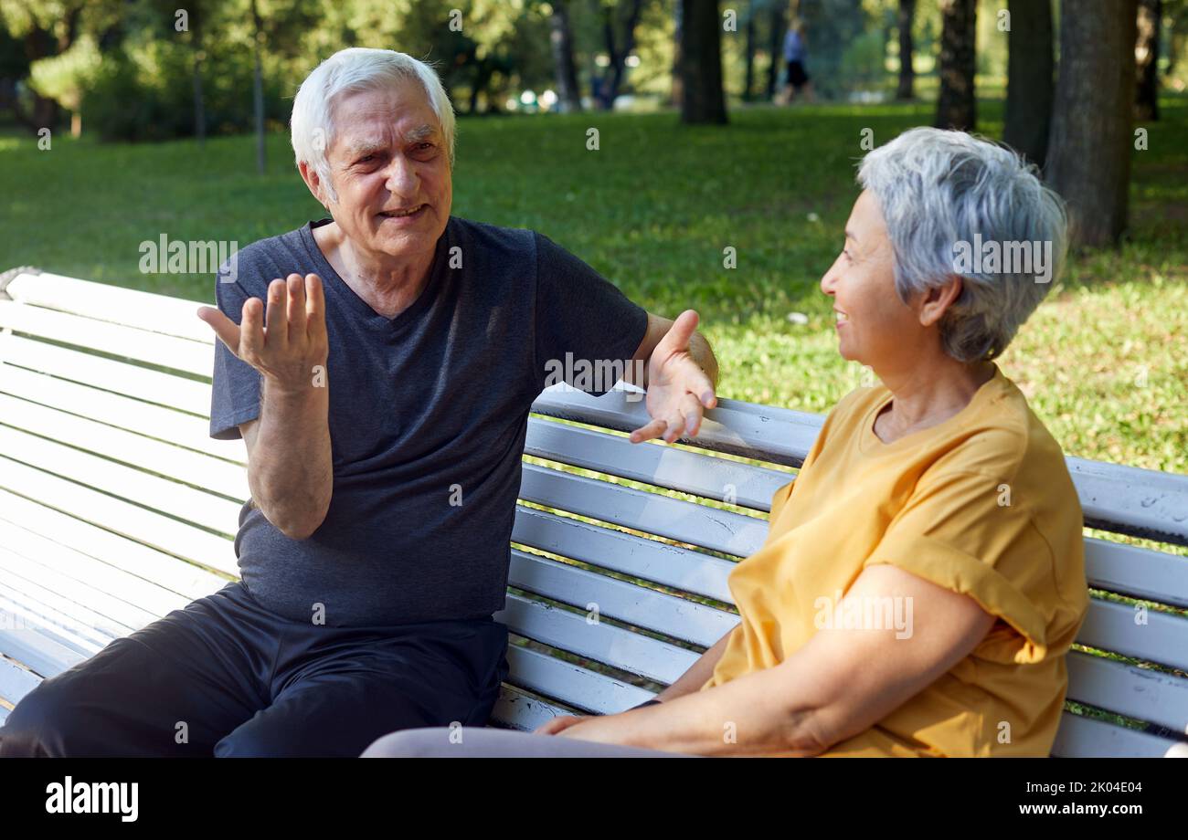 Older couple multi-racial spouses sit rest on bench enjoy talk in public summer park having friendly conversation share thoughts or news during meetin Stock Photo