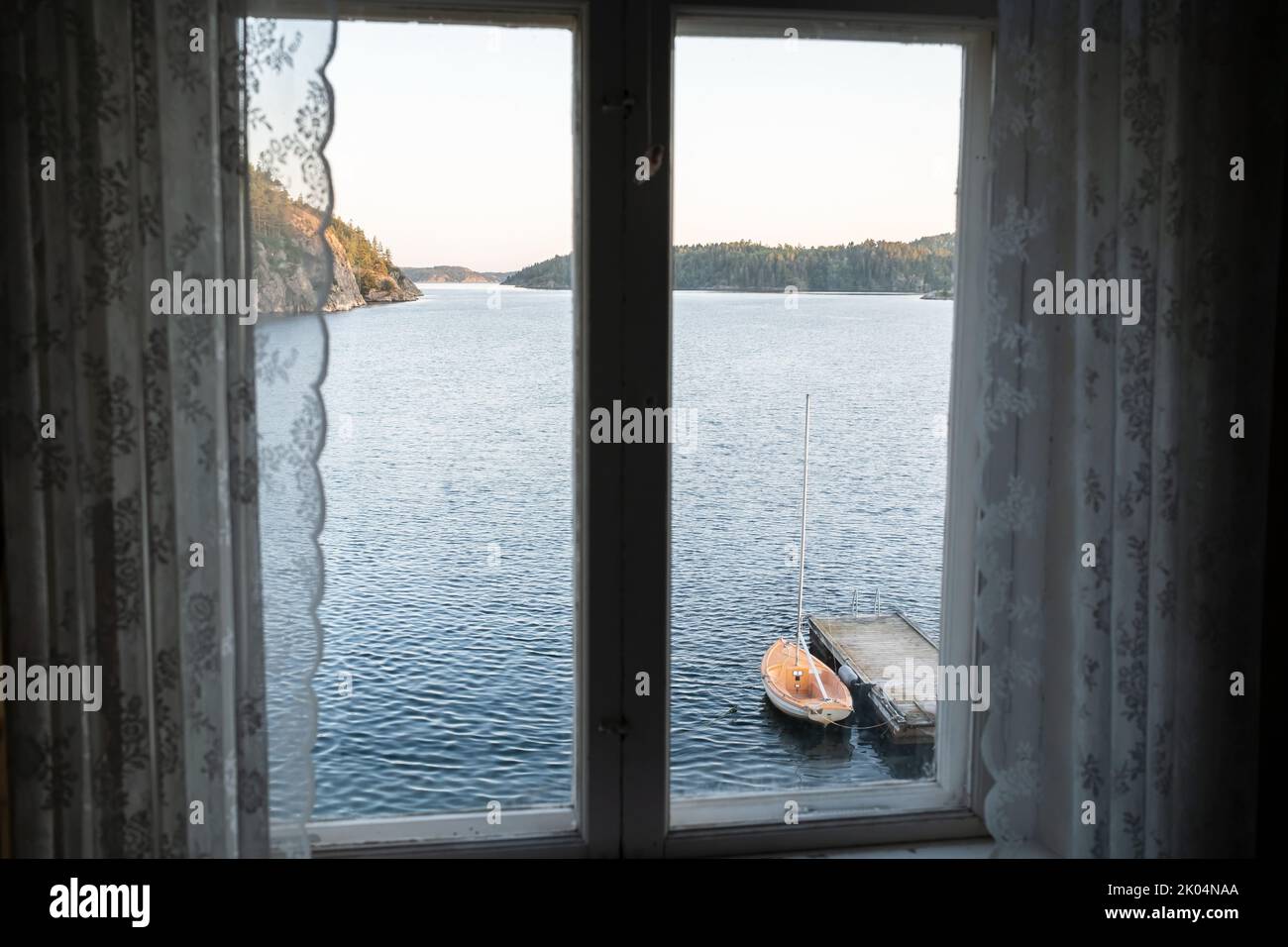 Old closed window across with great views of the sea, rocky shore, moored boat and sky at sunset.  Stock Photo